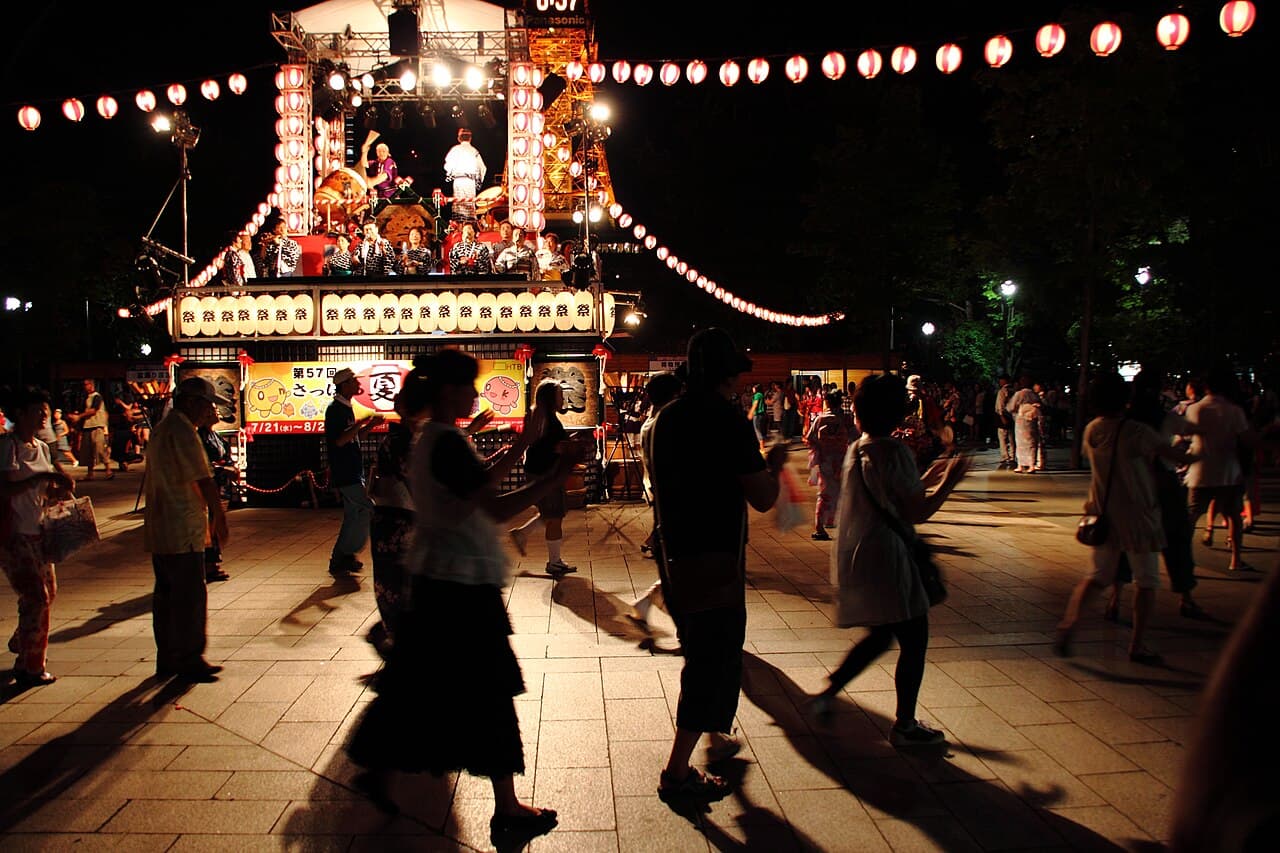 A lit yagura tower and strings of red paper lanterns at a Japanese summer festival at night, with a crowd