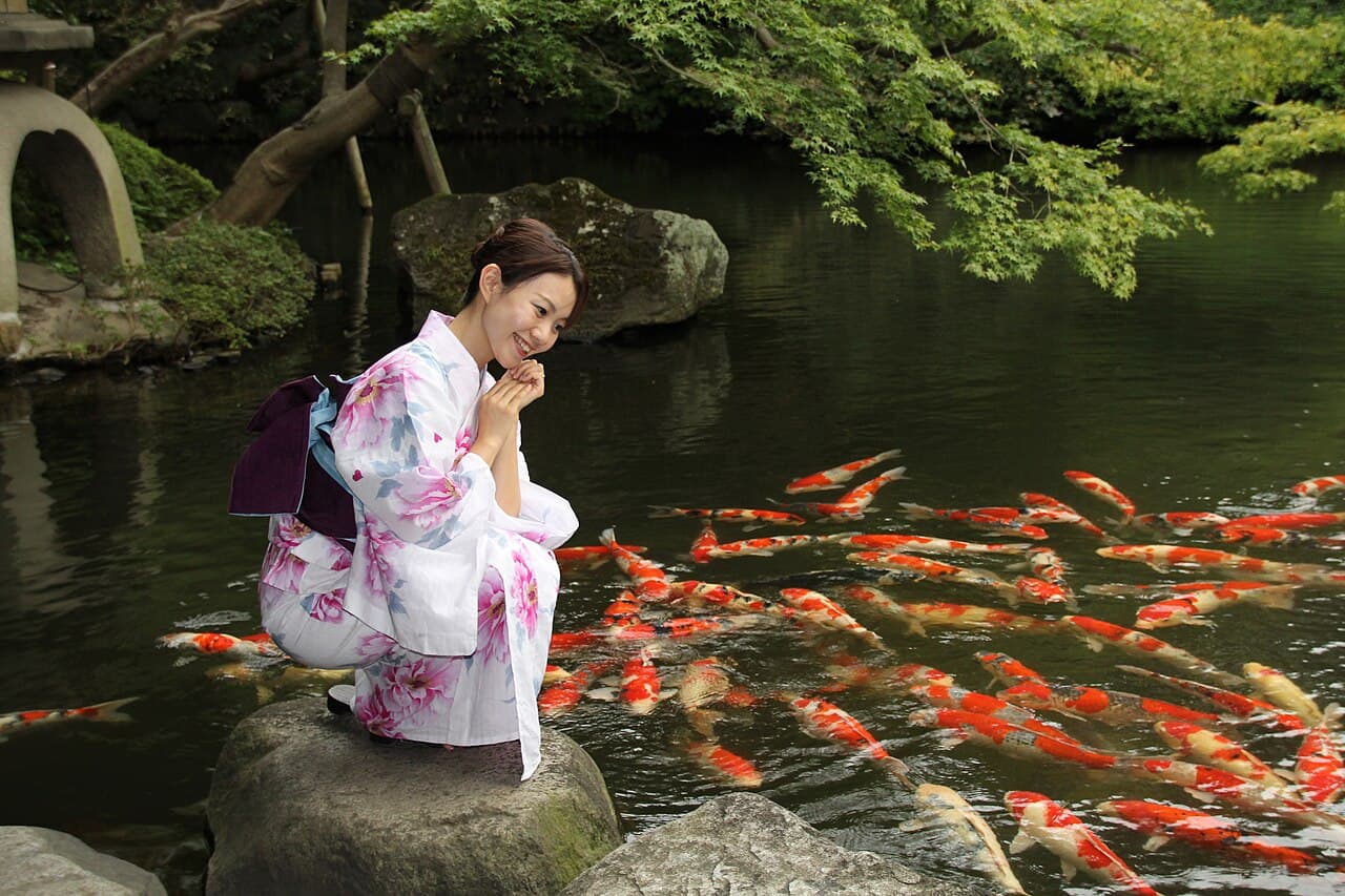 A woman wearing a patterned cotton yukata tied with an obi sash, standing by a koi pond in a Japanese garden